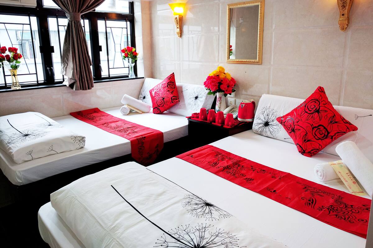 A hotel room with two beds, a window with curtains, a vase with flowers, and a wall-mounted mirror.