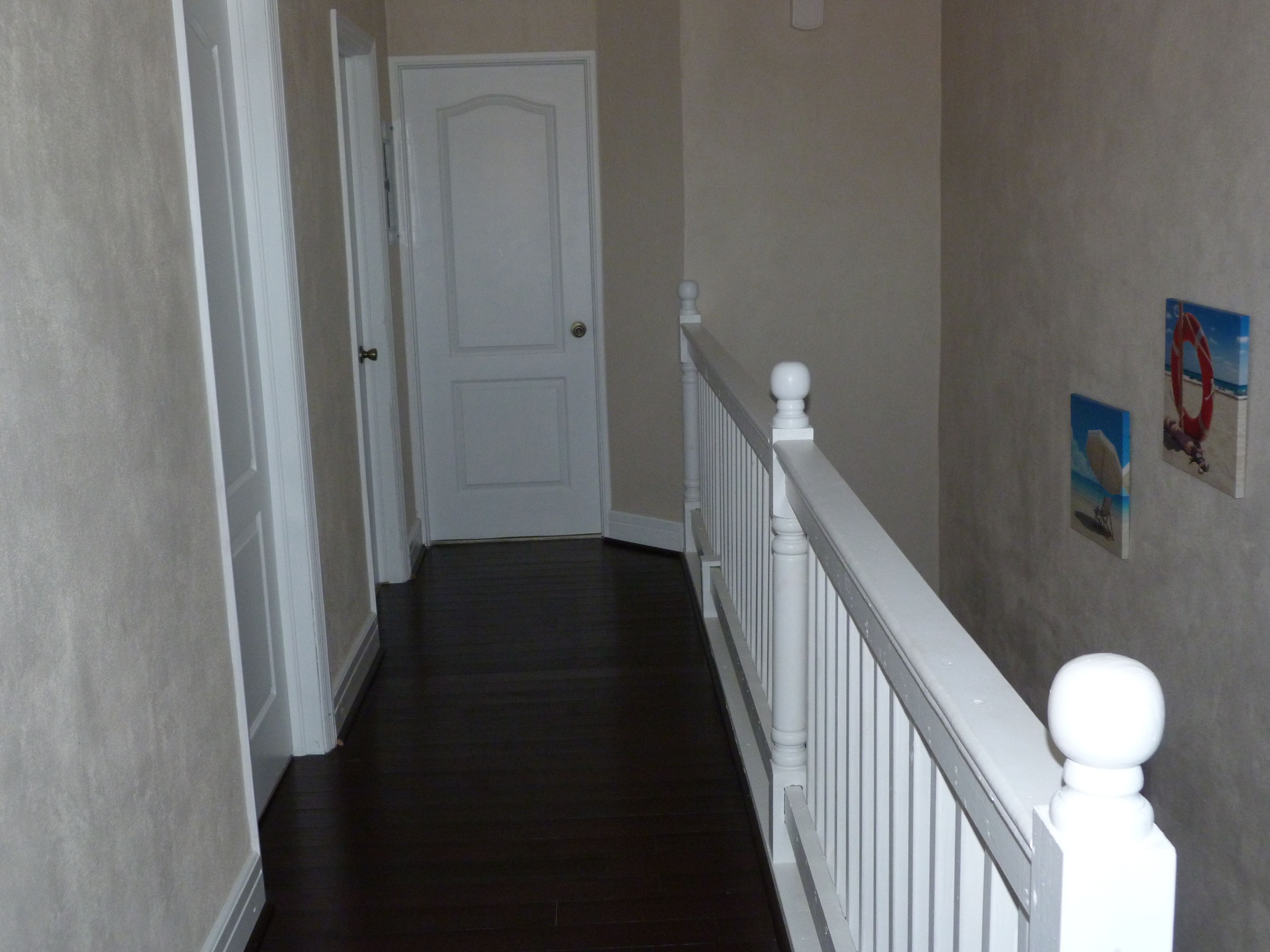A narrow hallway with a white door at the end, dark wooden flooring, and a white railing with a baluster.