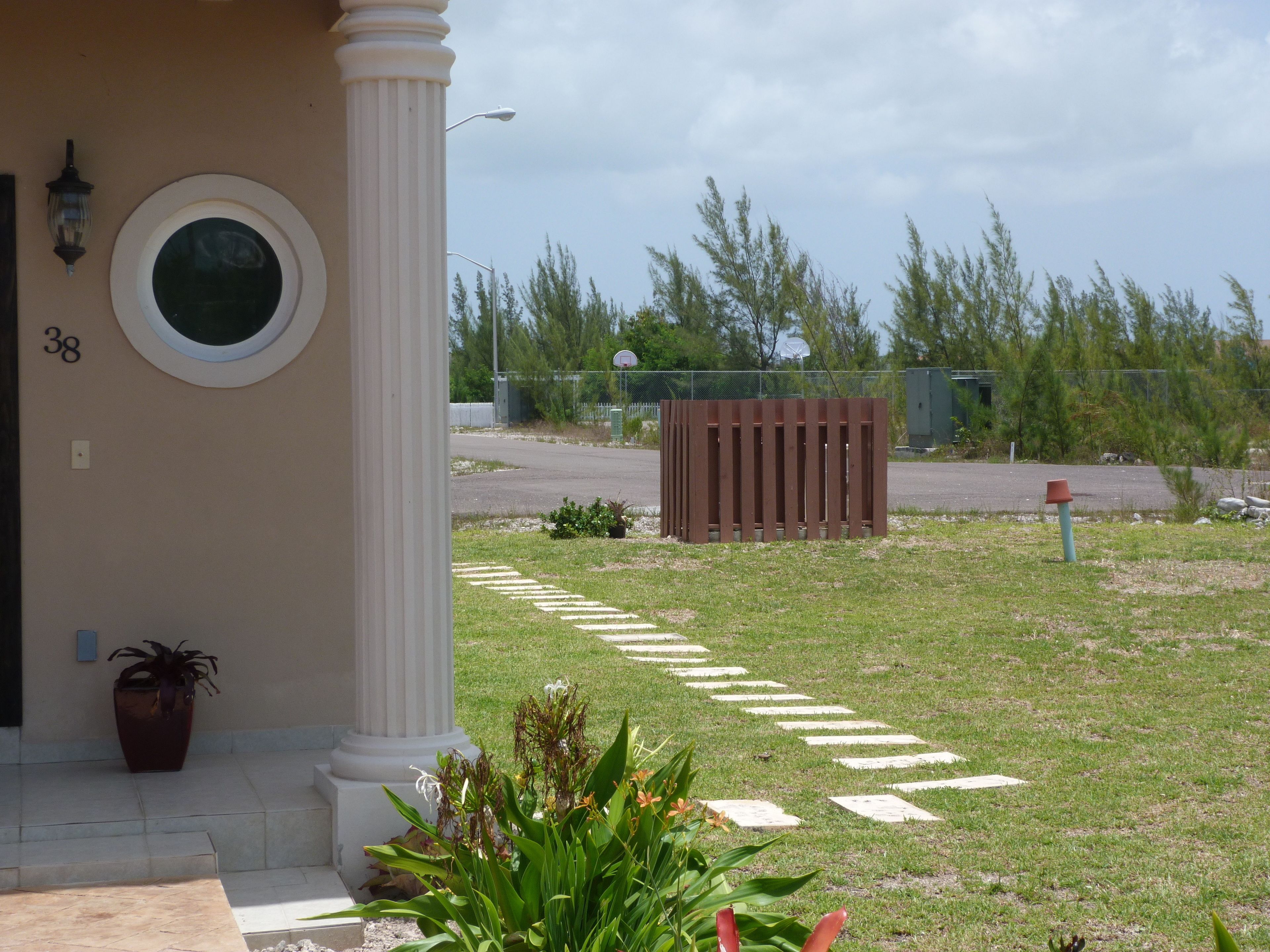A building entrance with a white column, a potted plant, and a paved pathway leading to a gate.