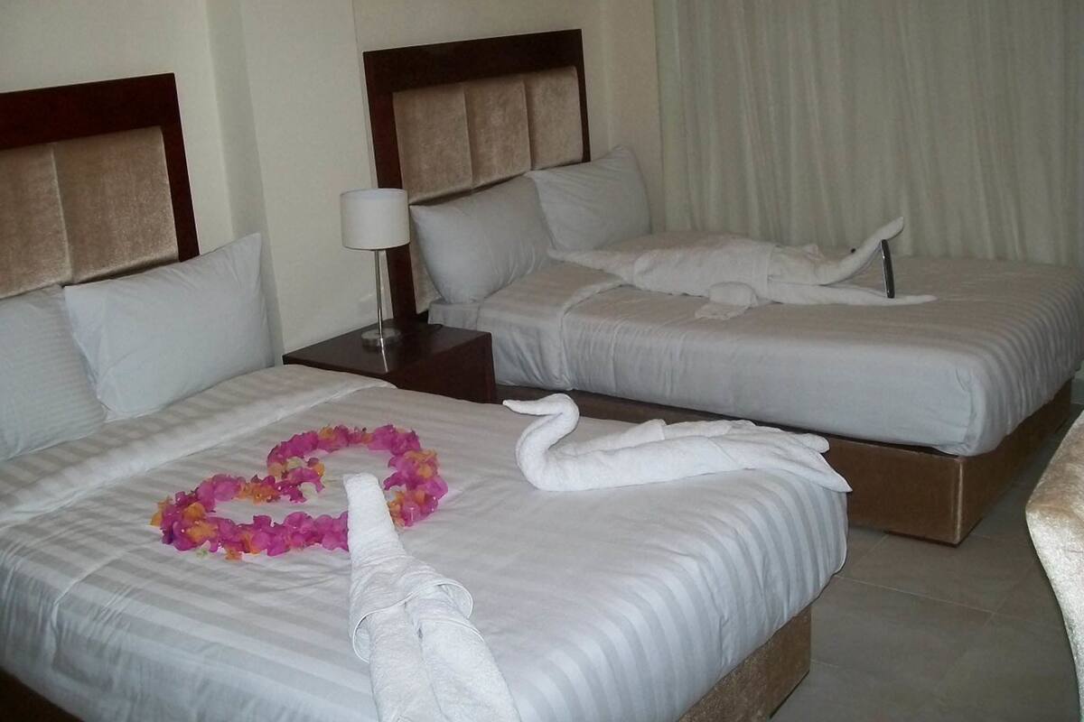 A hotel room with two beds, each with white linens and pillows, a nightstand with a lamp, and a decorative towel arrangement on one bed.