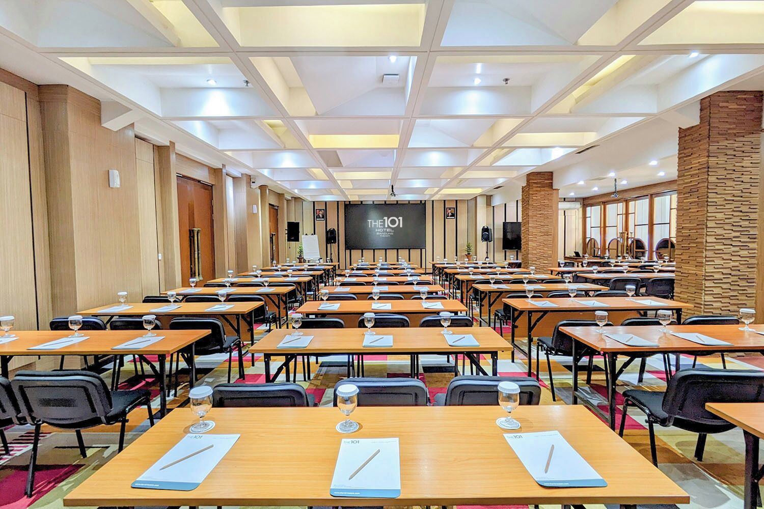 Un salón de conferencias con varias mesas y sillas, equipado con vasos de agua y blocs de notas, y una pantalla grande al frente.