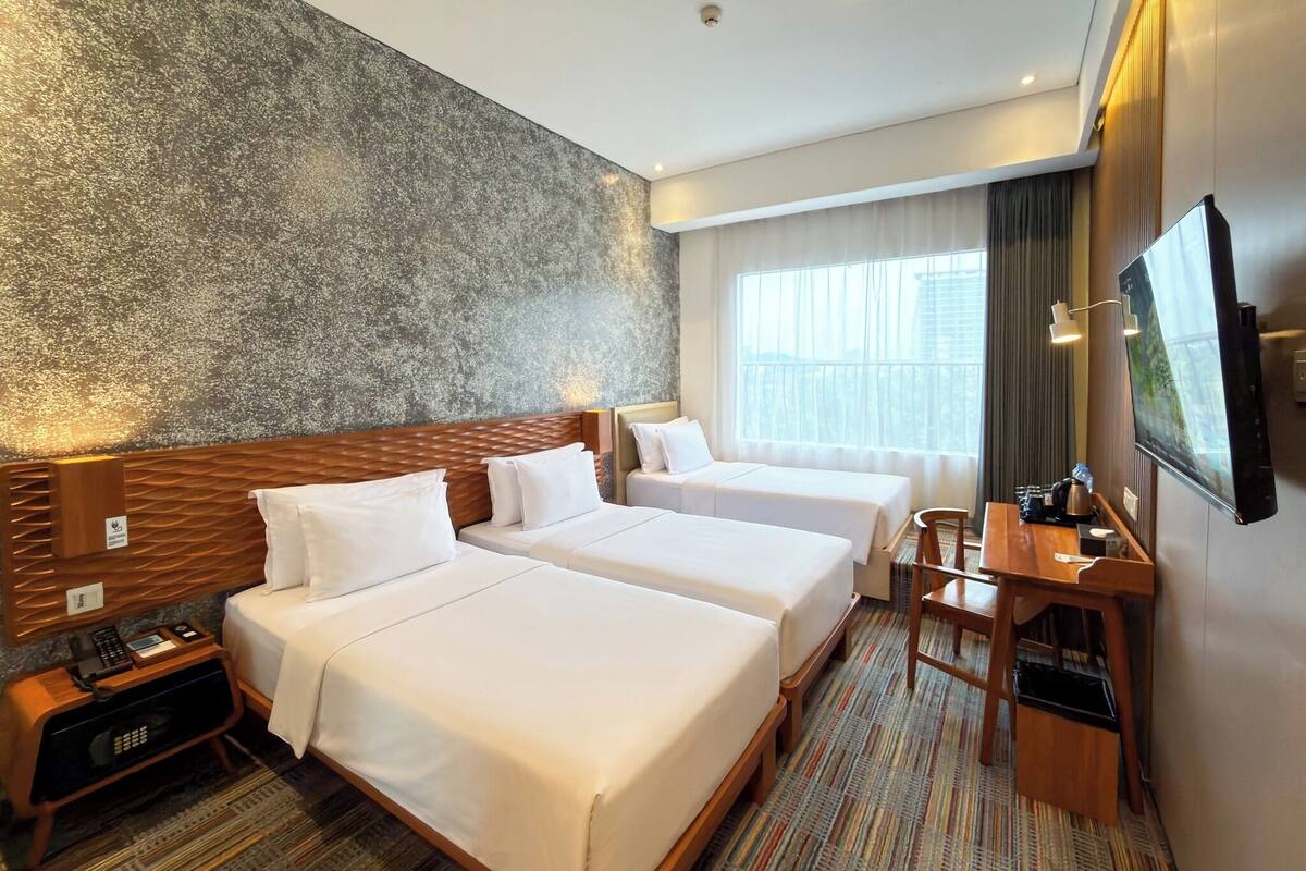 Quarto de hotel com duas camas, uma escrivaninha, uma cadeira, uma televisão e uma janela grande com cortinas.
