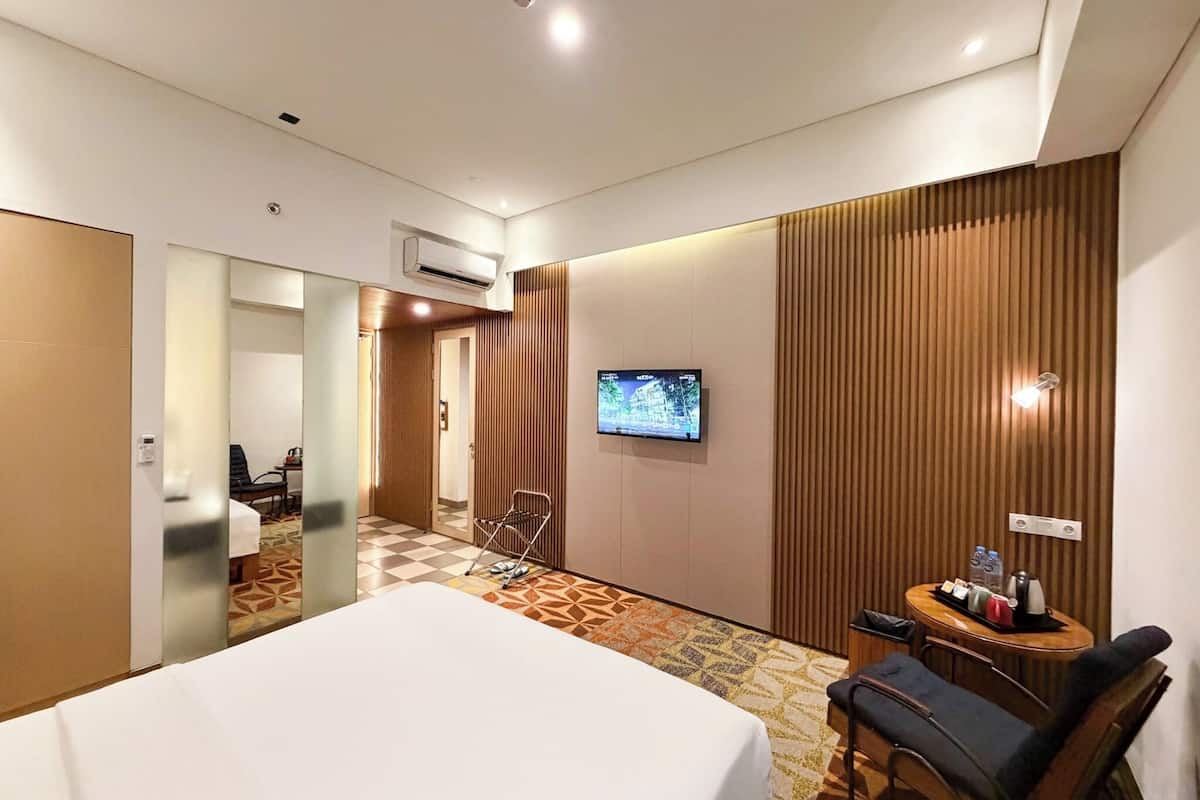 Quarto de hotel com cama, televisão, mesinha com bandeja de objetos, cadeira e ar condicionado fixado na parede.