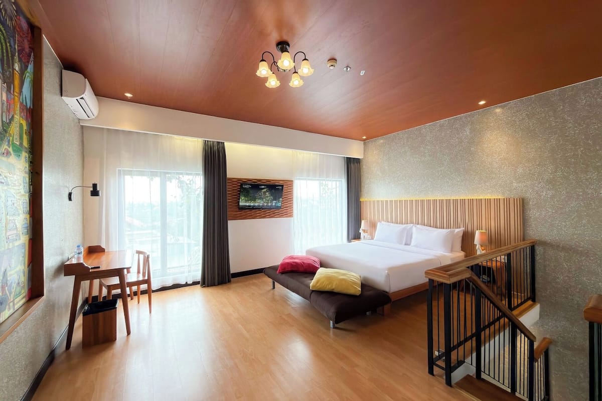 Quarto de hotel com cama, escrivaninha, cadeira, banco, escada e uma janela grande.