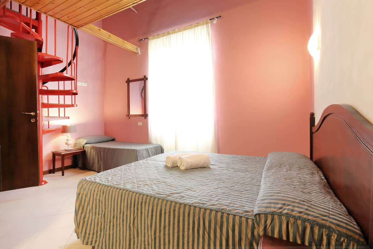 A bedroom with a bed, a nightstand, a lamp, a window with curtains, and a red spiral staircase.