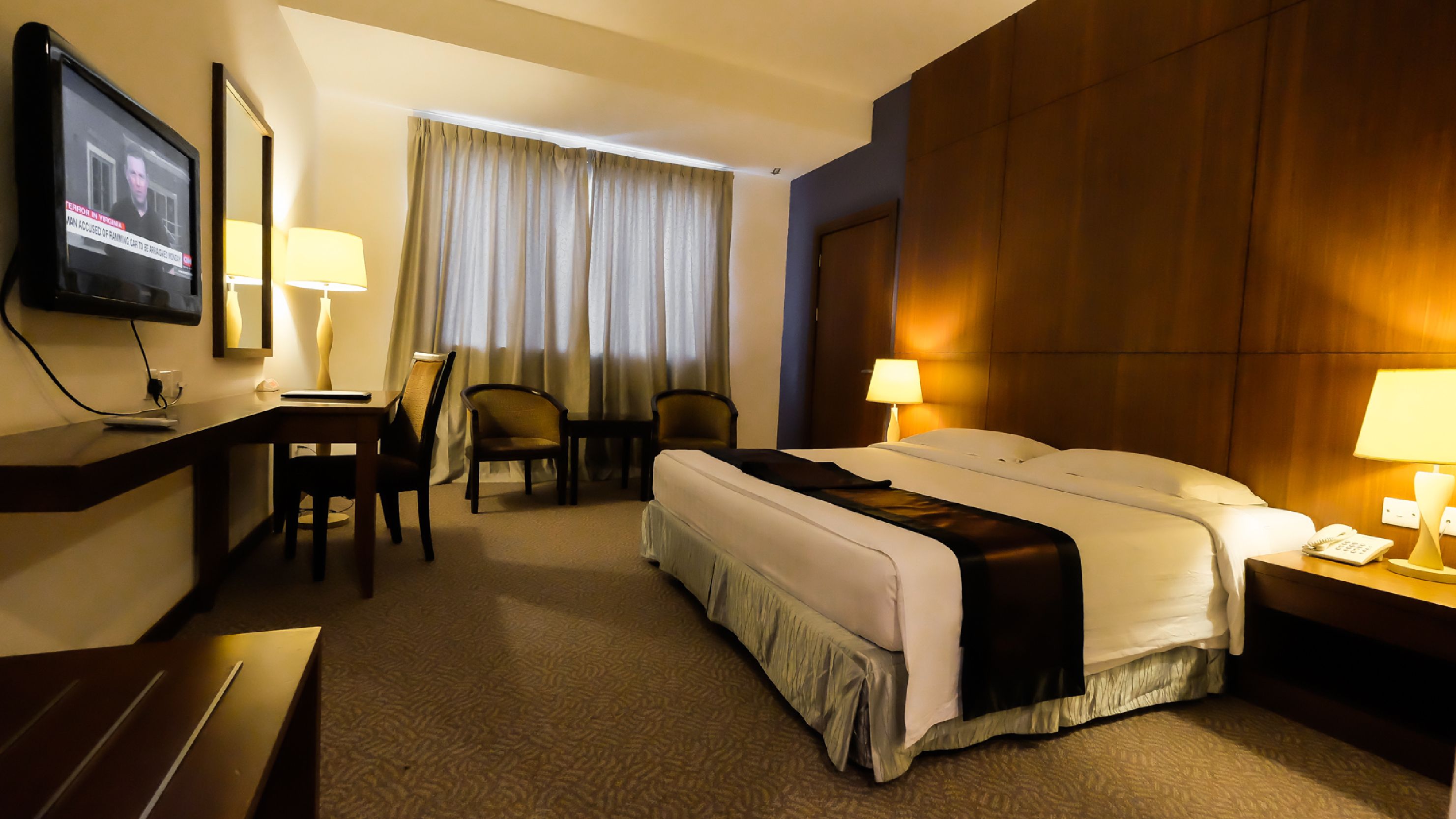 Una habitación de hotel con cama, dos sillas, escritorio, televisor y ventana con cortinas.