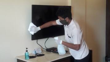 Una persona pulisce lo schermo di un televisore con un panno e una bottiglia spray, con una bottiglia di disinfettante per le mani e un telecomando su un tavolo.