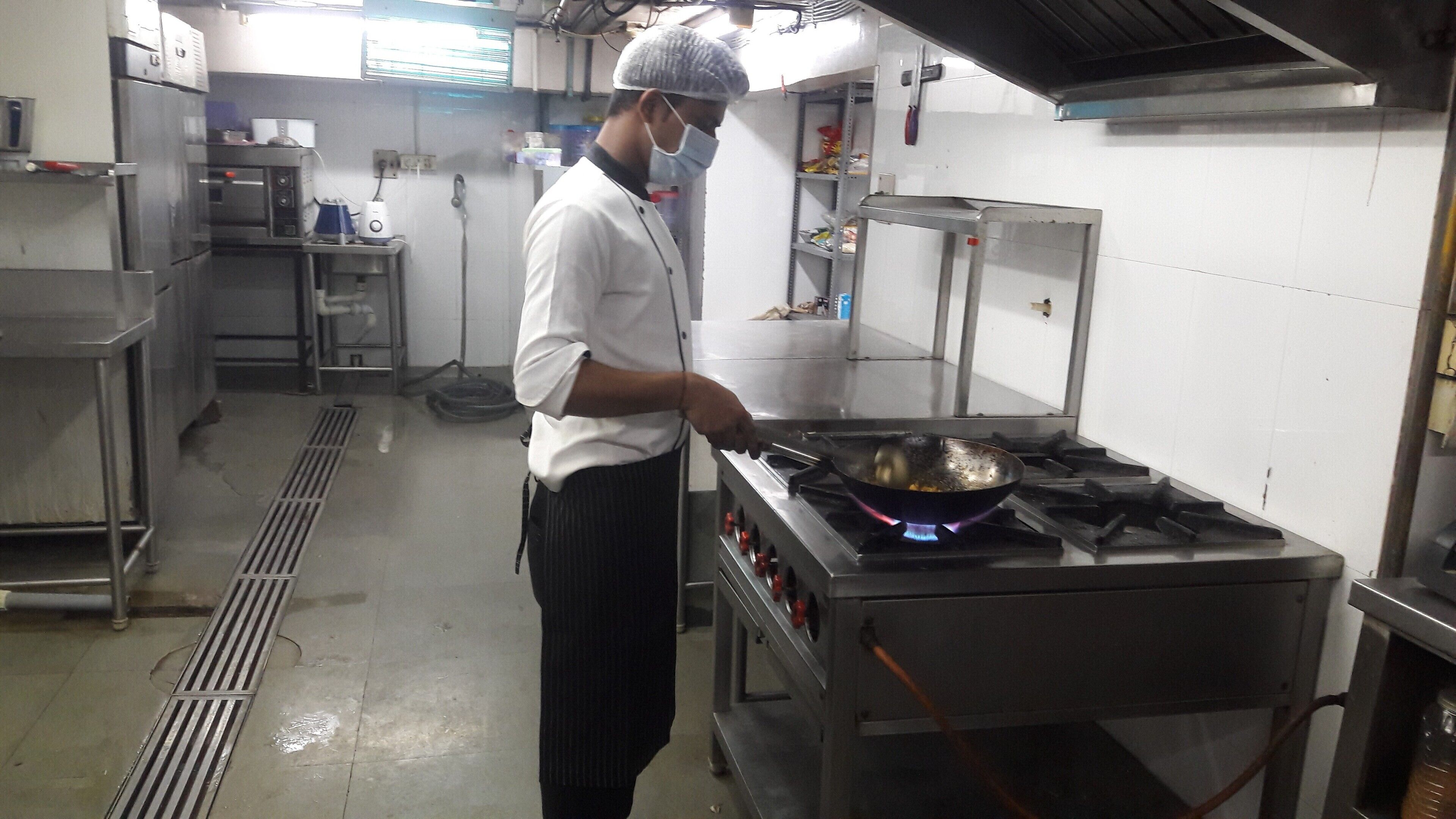 Un chef travaille dans une cuisine professionnelle équipée d’appareils en acier inoxydable, d’une cuisinière à gaz et d’une hotte aspirante.