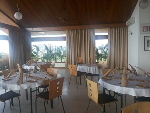 A dining area with tables set for two, wooden chairs, and a view of palm trees through large windows.