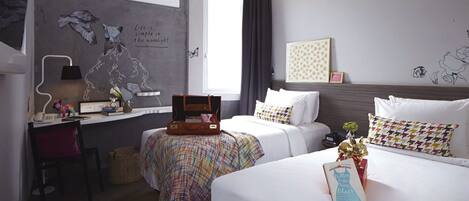 A hotel room with two beds, a desk, a chair, a window with curtains, and a wall with a mural and a framed picture.