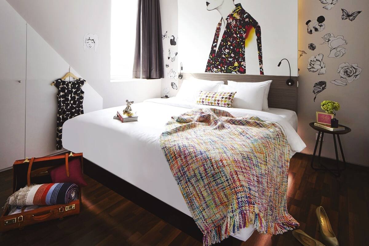 A modern bedroom with a large bed, a colorful blanket, a wall mural, and a small side table with a plant.