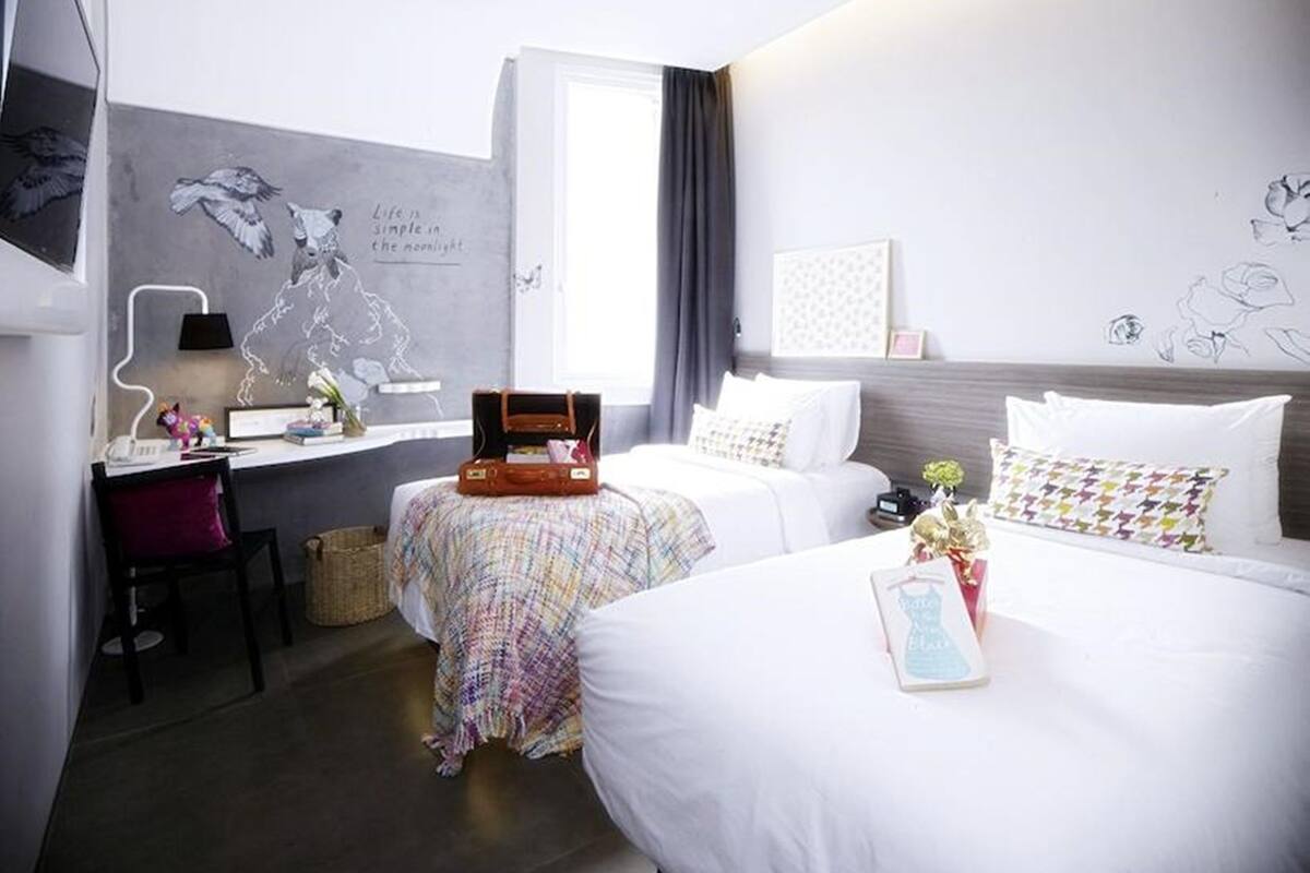 A hotel room with a bed, a desk, a chair, a suitcase, and a wall with a mural.
