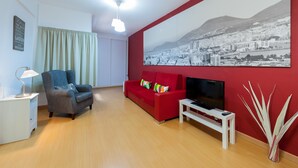 A living room with a red sofa, a blue armchair, a white side table, a TV, and a large cityscape painting on the wall. - Hotel Las Palmas Urban Center (Las Palmas de Gran Canaria)