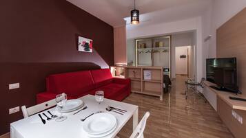 A modern living room with a red sofa, a dining table set with plates and cutlery, a television, and a kitchen area in the background.