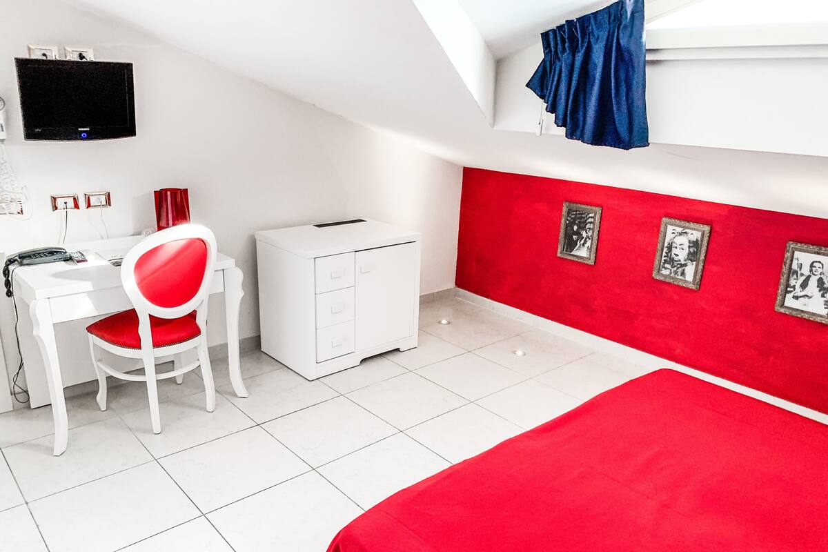 Ein Zimmer mit einem Bett, einem Schreibtisch mit Stuhl, einem kleinen weißen Schrank, einem an der Wand befestigten Fernseher und zwei gerahmten Bildern an der roten Wand.