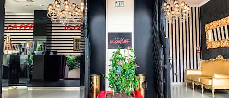 Un vestíbulo de hotel con alfombra roja, mostrador de recepción, candelabro, planta en maceta y sofá dorado.