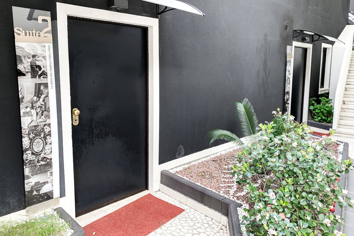 Eine schwarze Tür mit weißem Rahmen, eine kleine rote Fußmatte und ein Klingelknopf aus Metall. Vor der Tür befindet sich ein kleiner Garten mit Kies und einer Pflanze mit grünen Blättern und weißen Blüten.