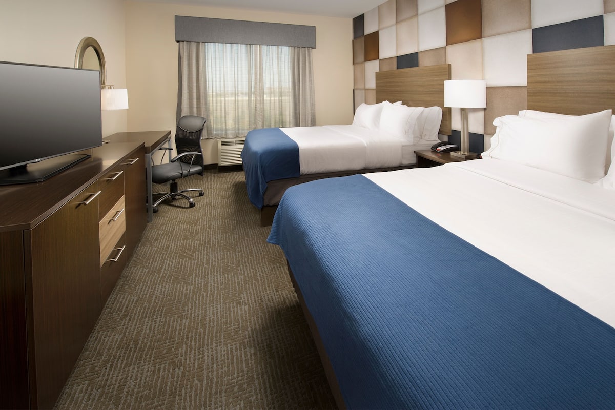 A hotel room with two beds, a desk with a TV, a chair, and a large window with curtains.