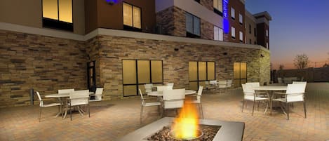Un patio al aire libre con varias mesas y sillas, una chimenea central y un edificio con fachada de piedra y ventanas.