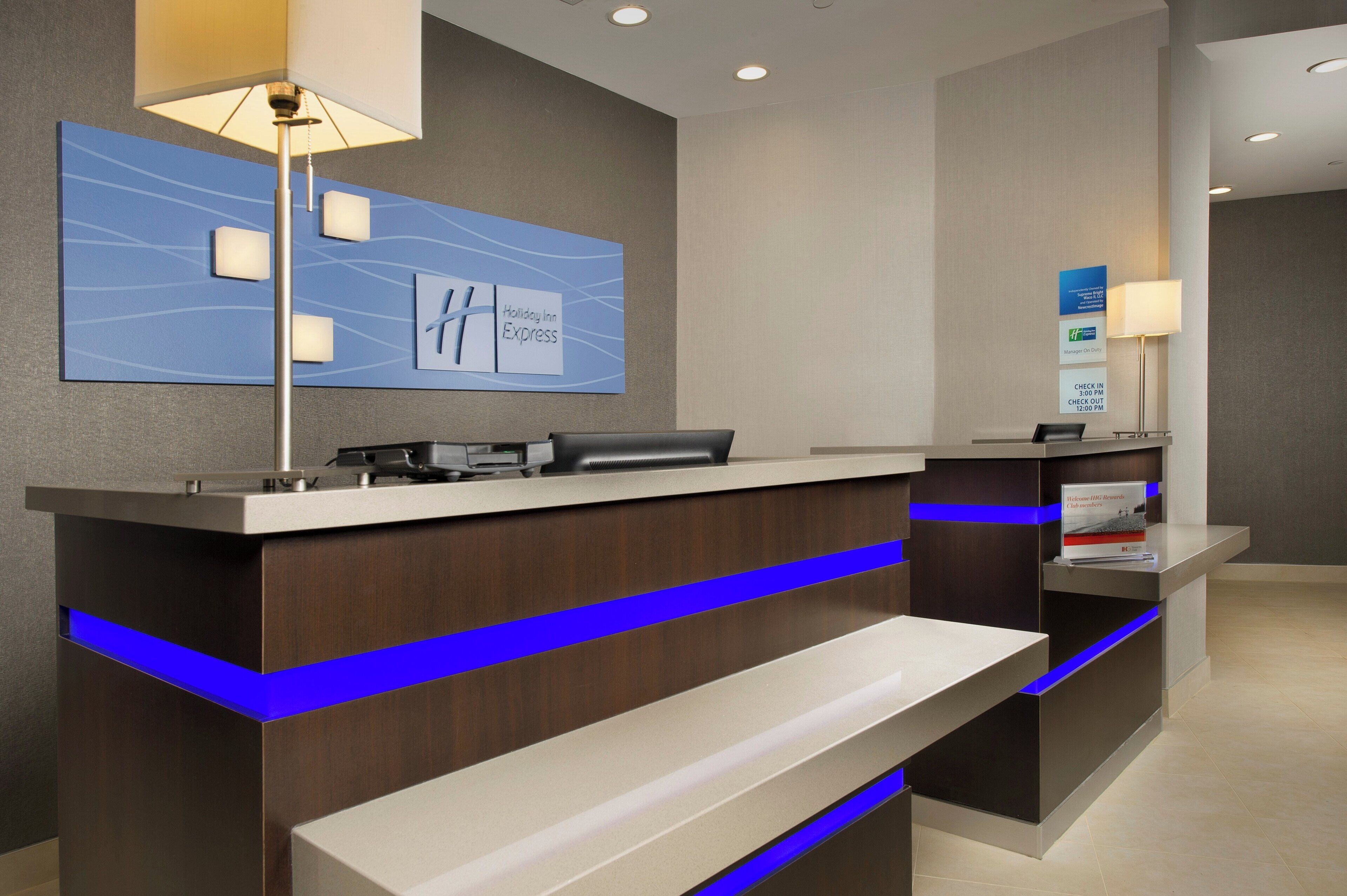 A modern hotel reception desk with a blue accent, a digital check-in kiosk, and a wall-mounted sign for the Holiday Inn Express.