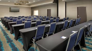 A conference room with rows of tables and chairs, a flat-screen TV on the wall, and a carpeted floor.