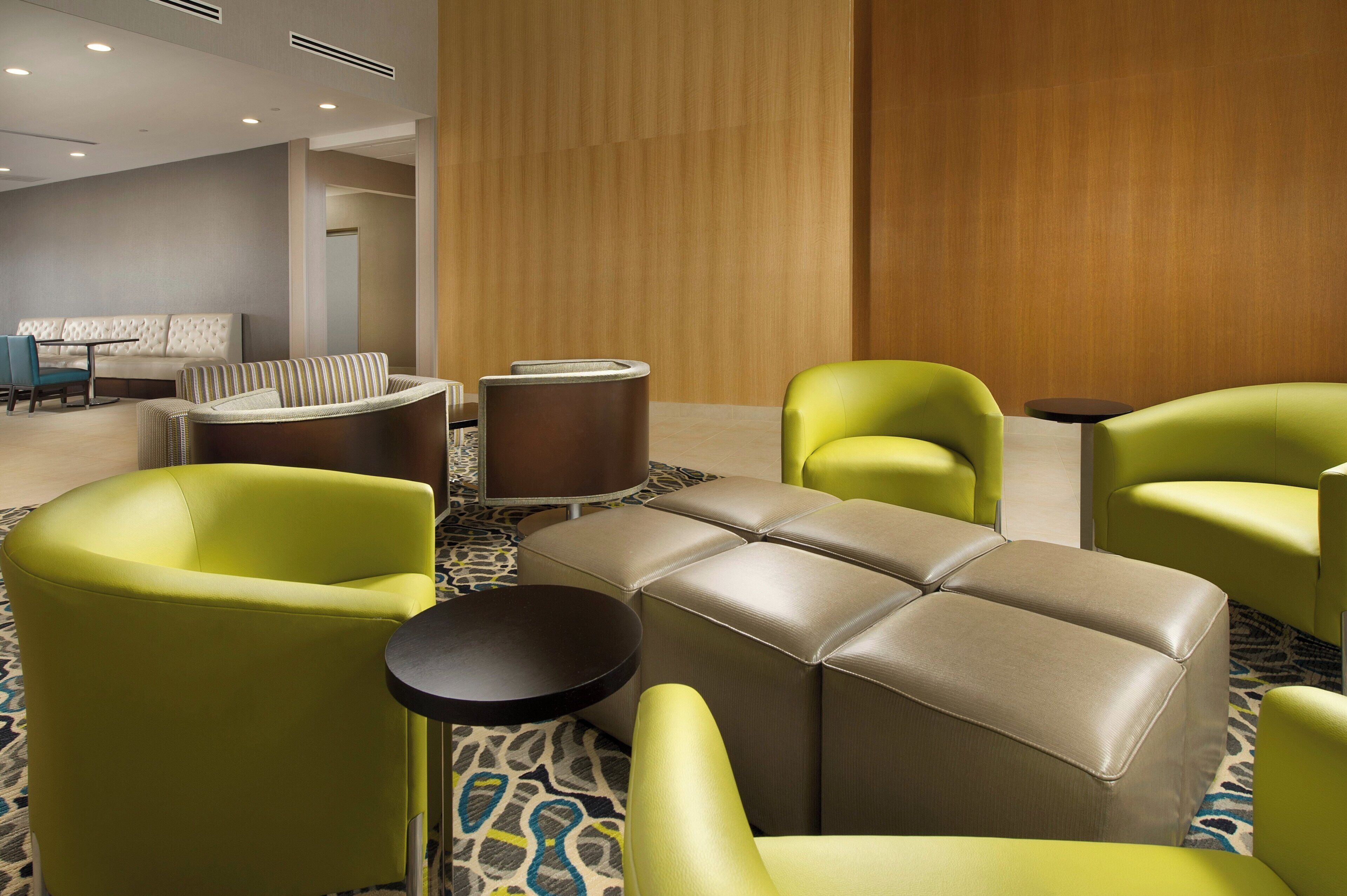 A hotel lobby with a seating area featuring a tufted sofa, a striped armchair, and two green armchairs. There is a round table and a small black table. The floor has a patterned carpet.