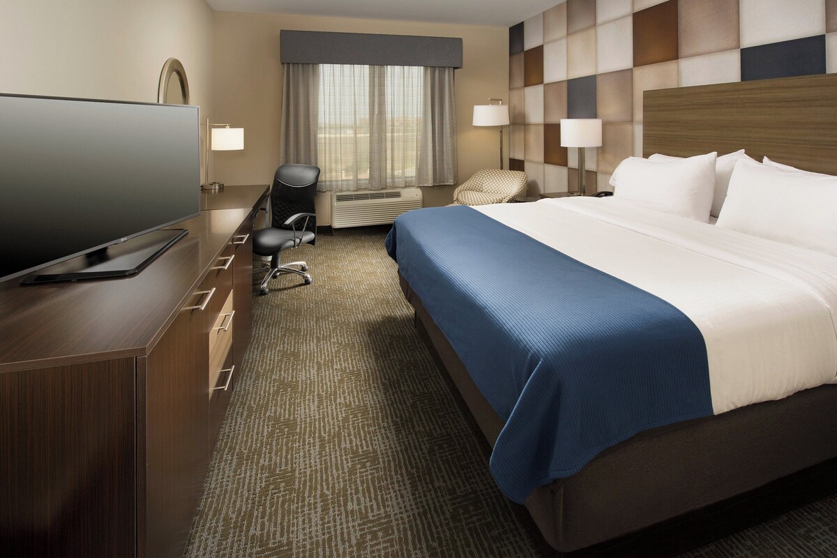 A hotel room with a large flat-screen TV, a bed with a blue bedspread, a desk with a computer, a chair, and a window with curtains.