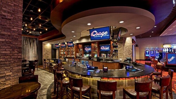 Zona de bar con barra circular, varios arreglos de asientos y un llamativo letrero de 'Cosage Casino Hotel'.