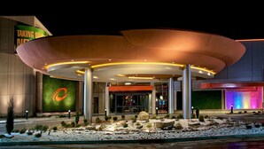 Una entrada de hotel moderna con techo curvo, un logotipo destacado en la pared y un área ajardinada con rocas y arbustos.