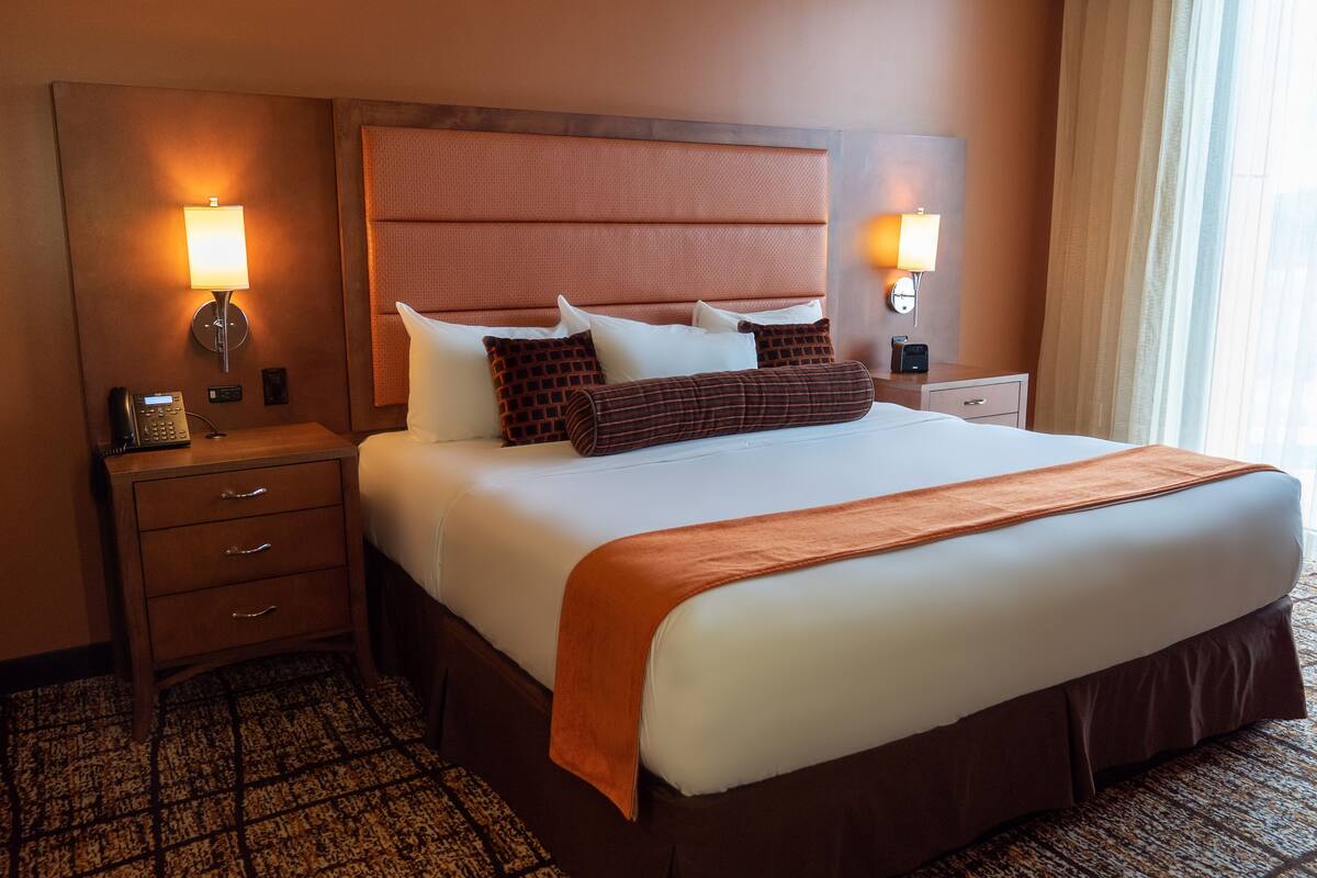 A hotel room with a large bed, two bedside tables, a nightstand, a wall-mounted lamp, and a patterned carpet.
