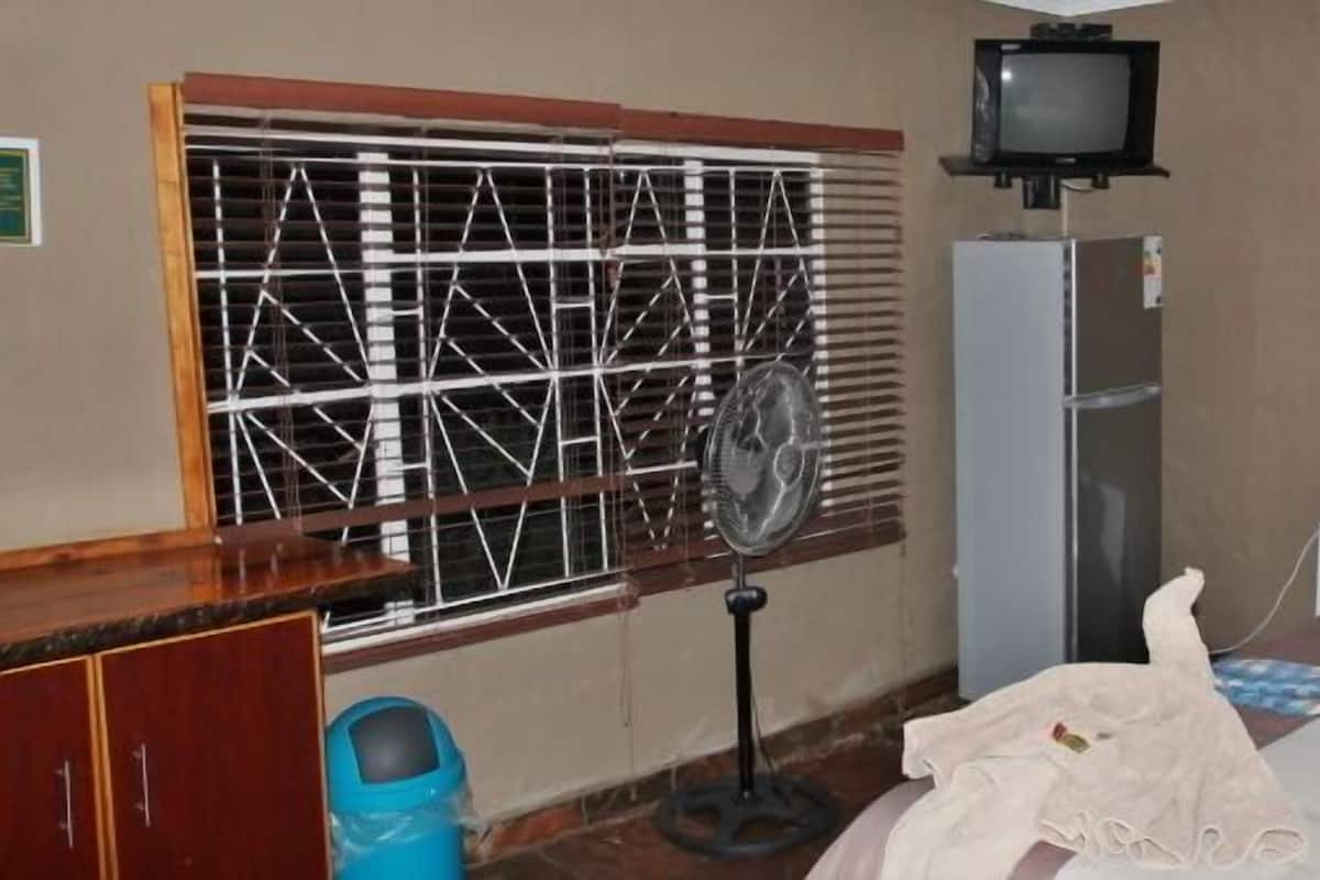 A room with a window with metal bars, a television on a stand, a refrigerator, a bed with a blanket and a pillow, and a wall with a framed picture.