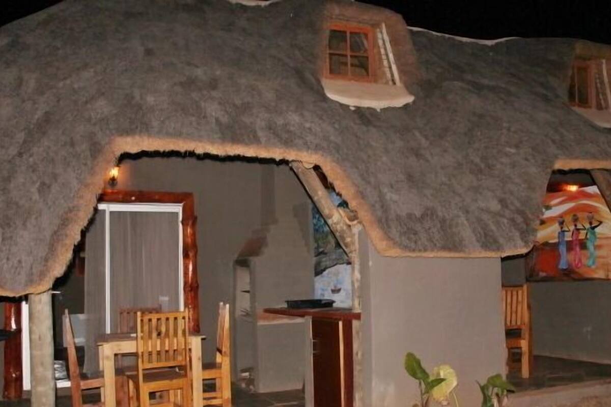 A thatched-roof hut with a small window and a door, a wooden table, and a chair outside.
