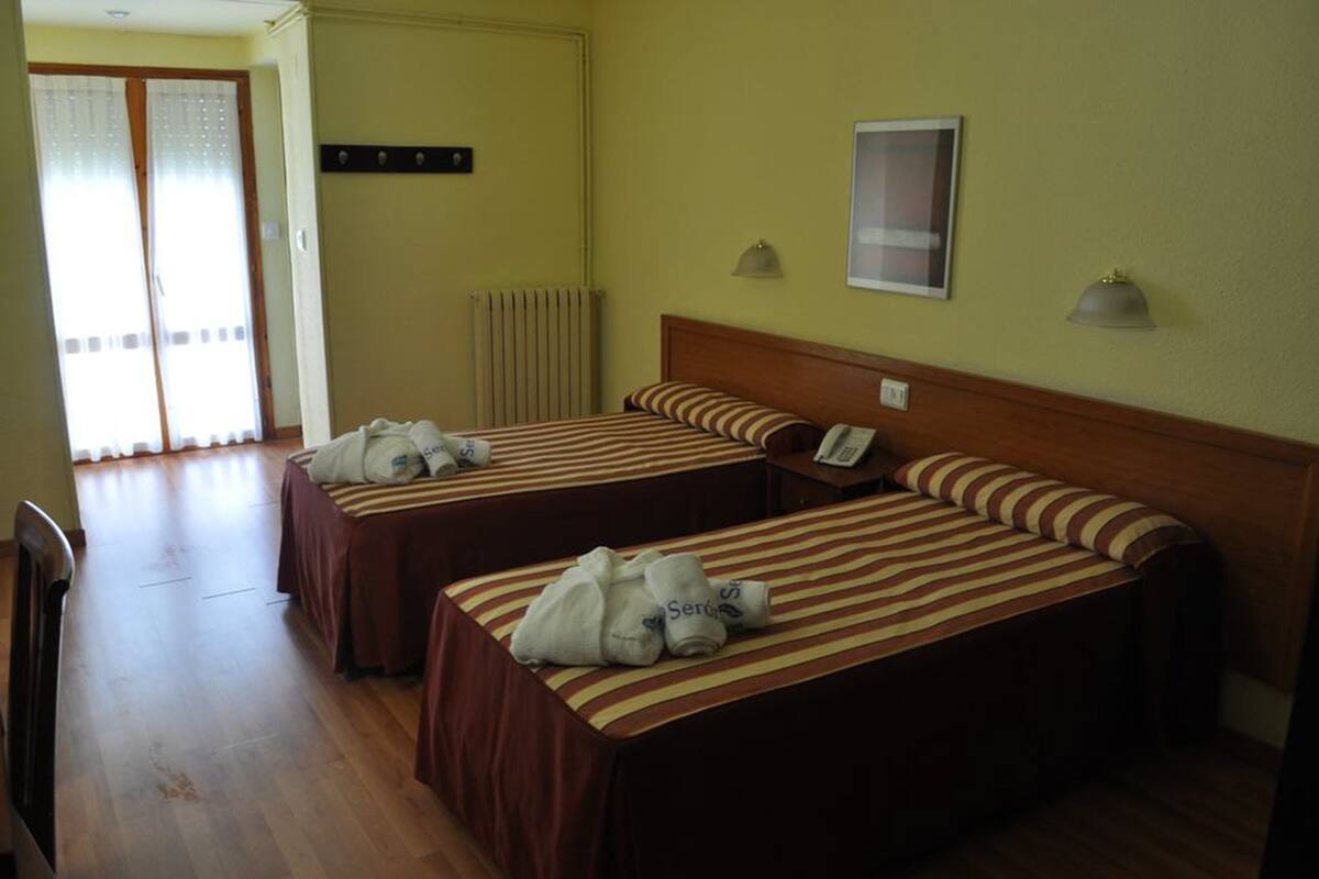 A hotel room with two beds, a wooden floor, a radiator, and a telephone on the wall.