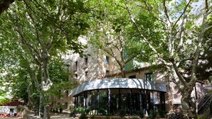 A covered outdoor seating area with a curved roof, surrounded by trees and a building with multiple windows. - Balneario Serón (Jaraba)