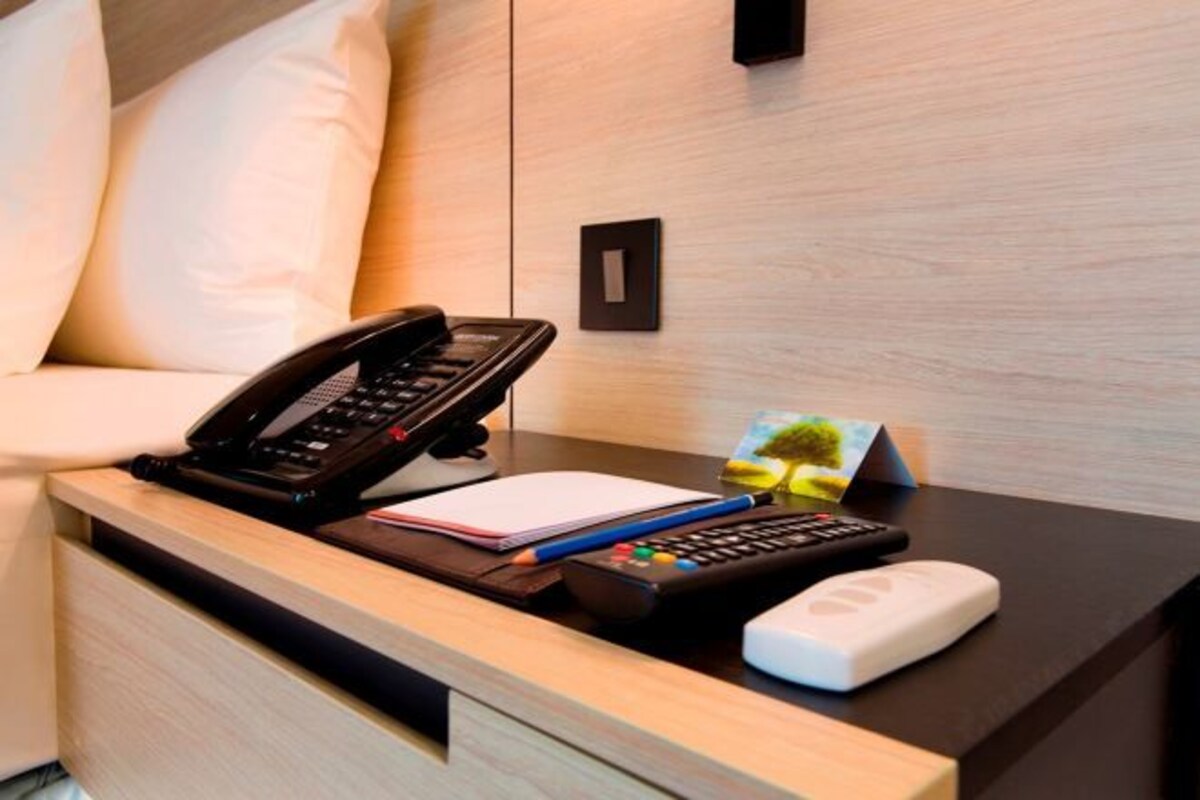 Hotelkamer met een houten nachtkastje, telefoon, notitieboekje, pen, afstandsbediening en een wit apparaat, mogelijk een thermostaat of bedieningspaneel.