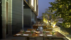 Un espace extérieur aménagé pour les repas, avec des tables et des chaises, une rambarde en verre et la façade d’un bâtiment en arrière-plan.