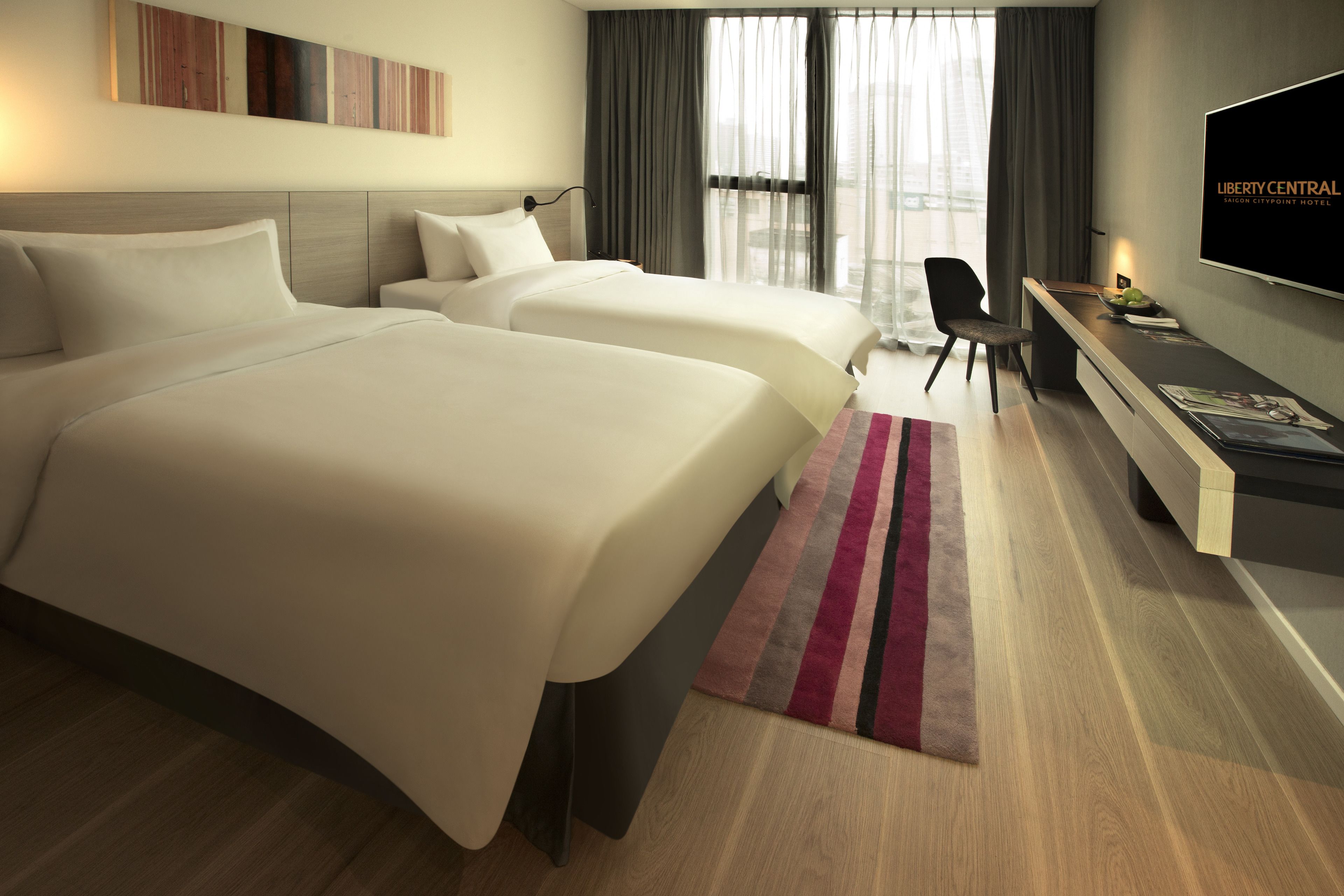 A hotel room with two beds, a desk, a TV, and a view of the city.
