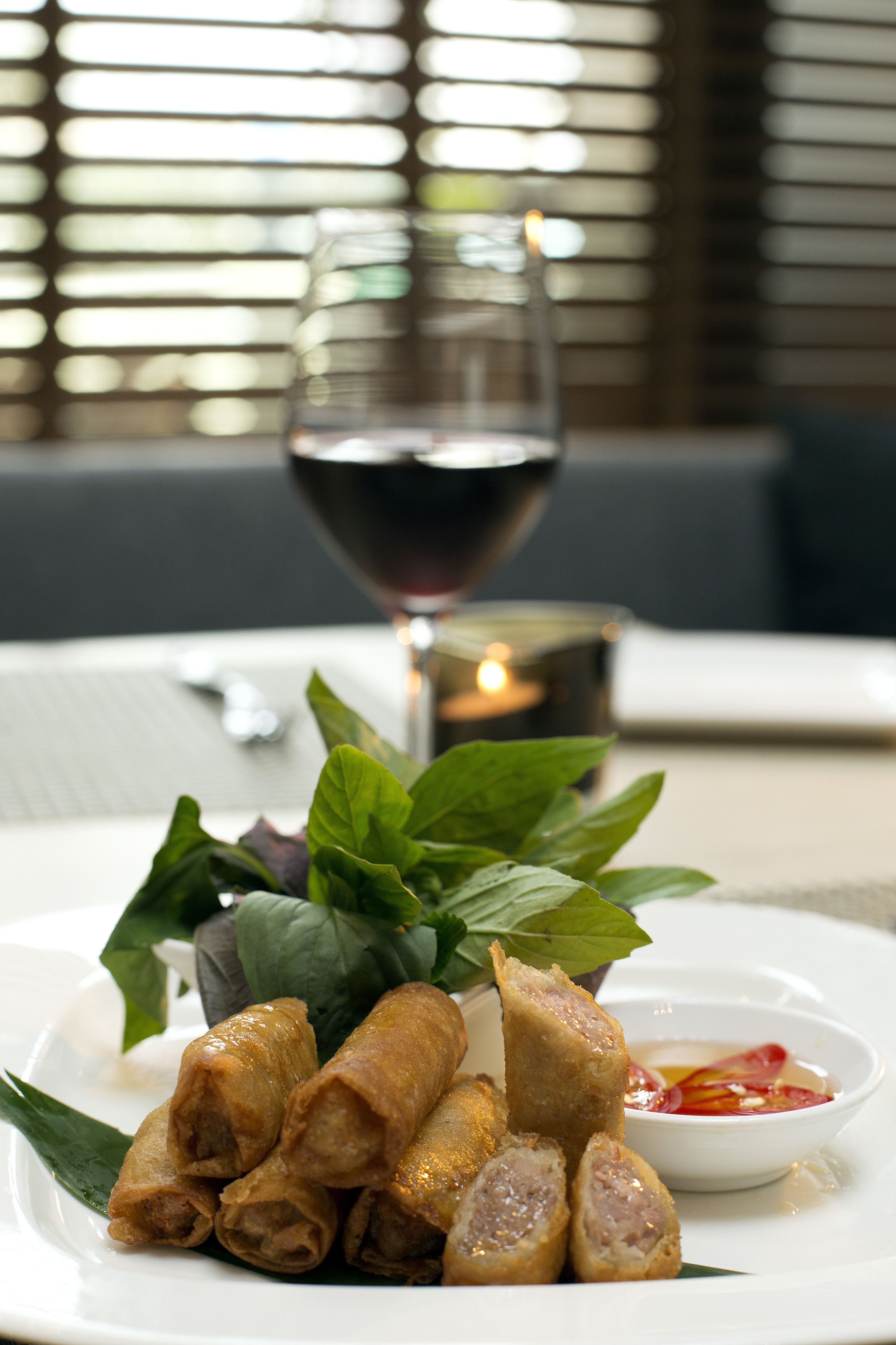 A plate of spring rolls with a dipping sauce, a glass of red wine, and a candle on a table.