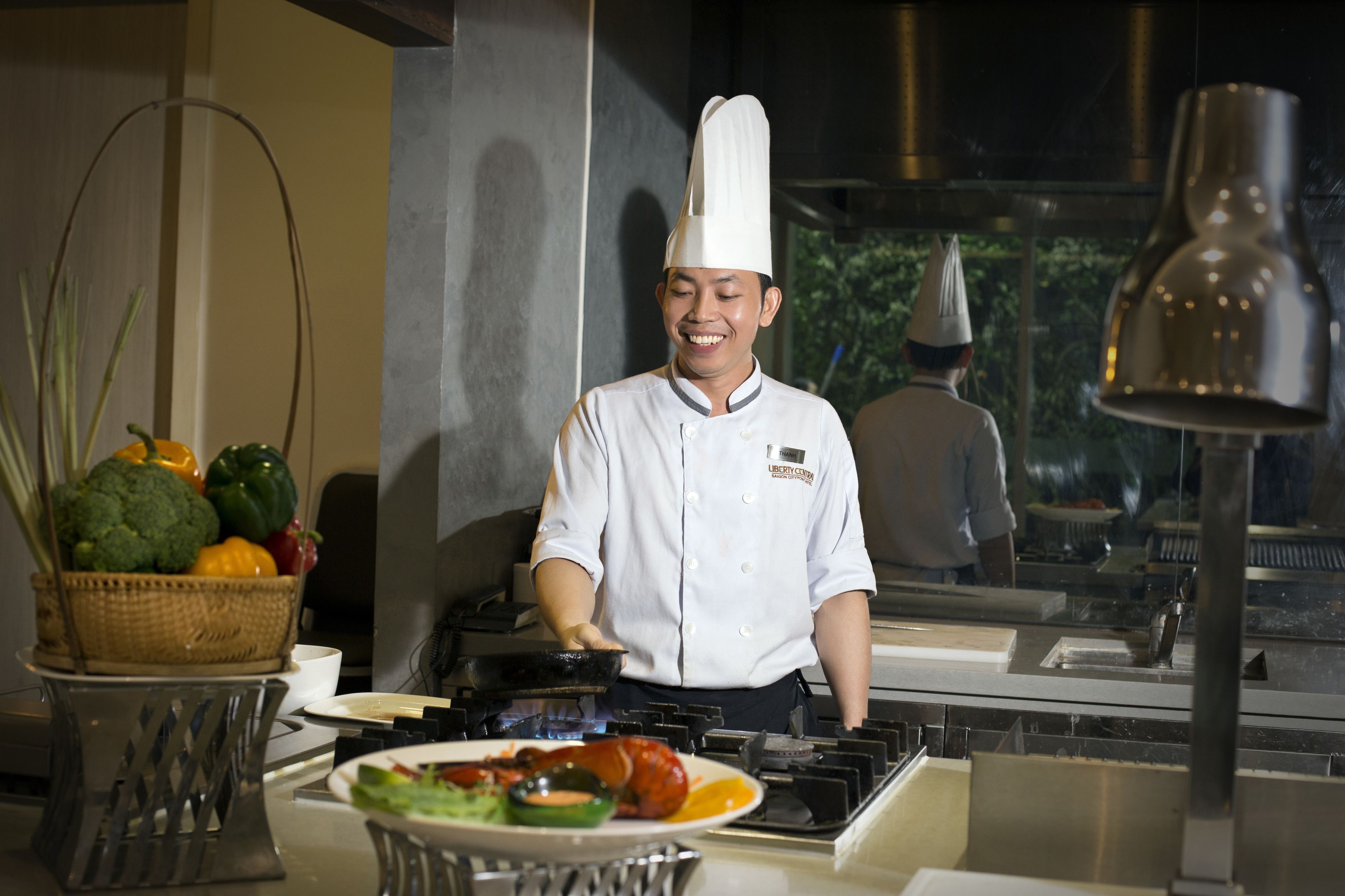 A chef in a white uniform and hat cooking in a professional kitchen with stainless steel appliances and a view of greenery outside.