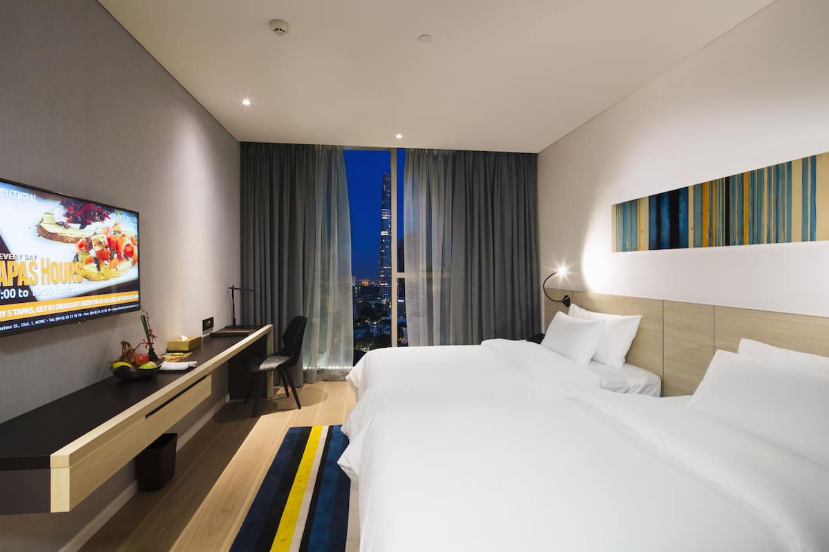Ein Hotelzimmer mit Bett, Schreibtisch, Fernseher und Blick auf eine nächtliche Stadtlandschaft.