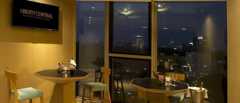 A modern hotel room with a city view, a round dining table, and two bar stools.