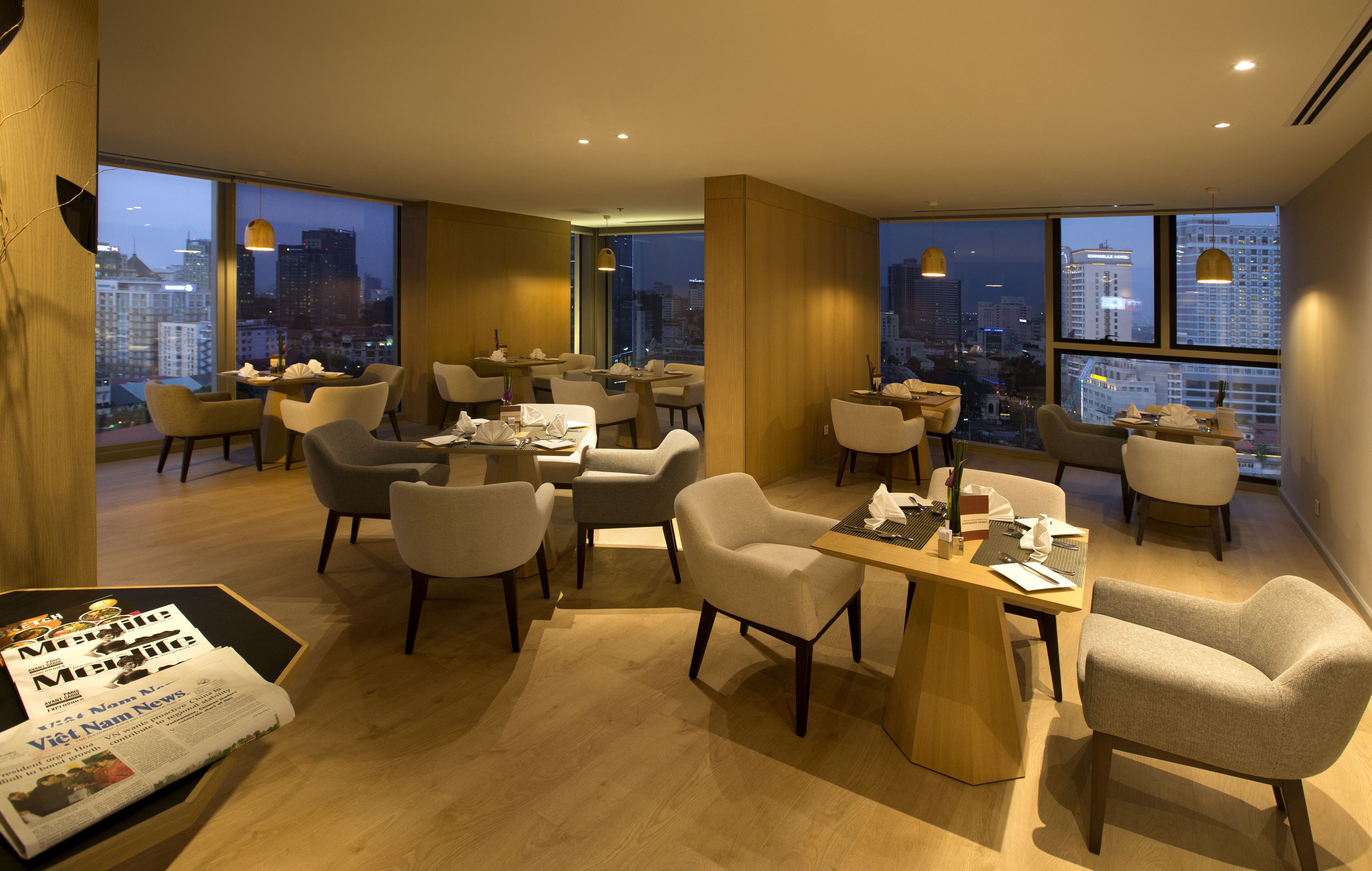 A modern hotel lobby with multiple seating areas, large windows offering city views, and a newspaper on a table.