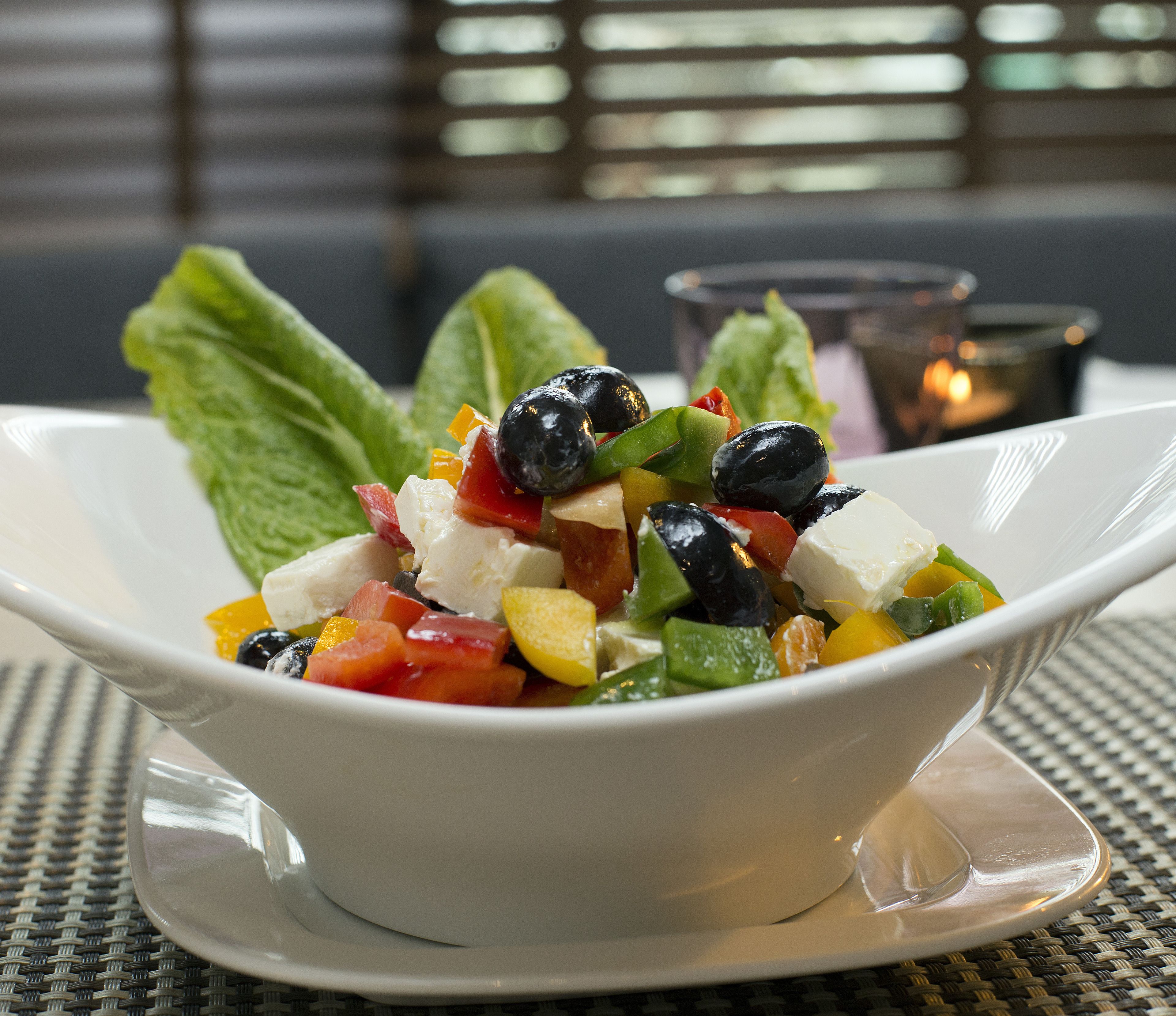 Un tazón blanco de ensalada con diversas verduras y aceitunas sobre un tapete tejido.