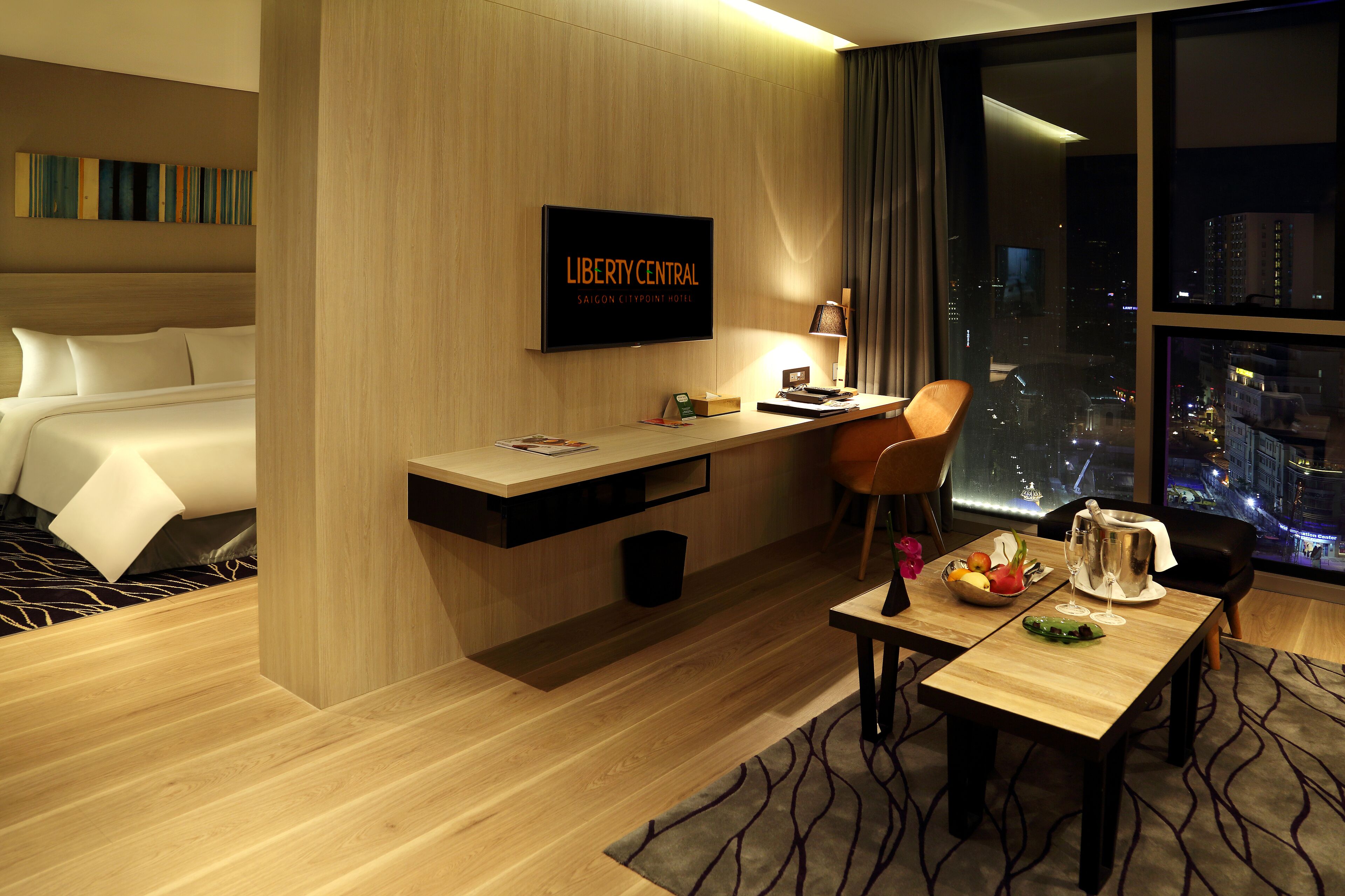 A hotel room with a bed, desk, and dining area. A television displays "Liberty Central Saigon Citypoint Hotel". The room has a city view.