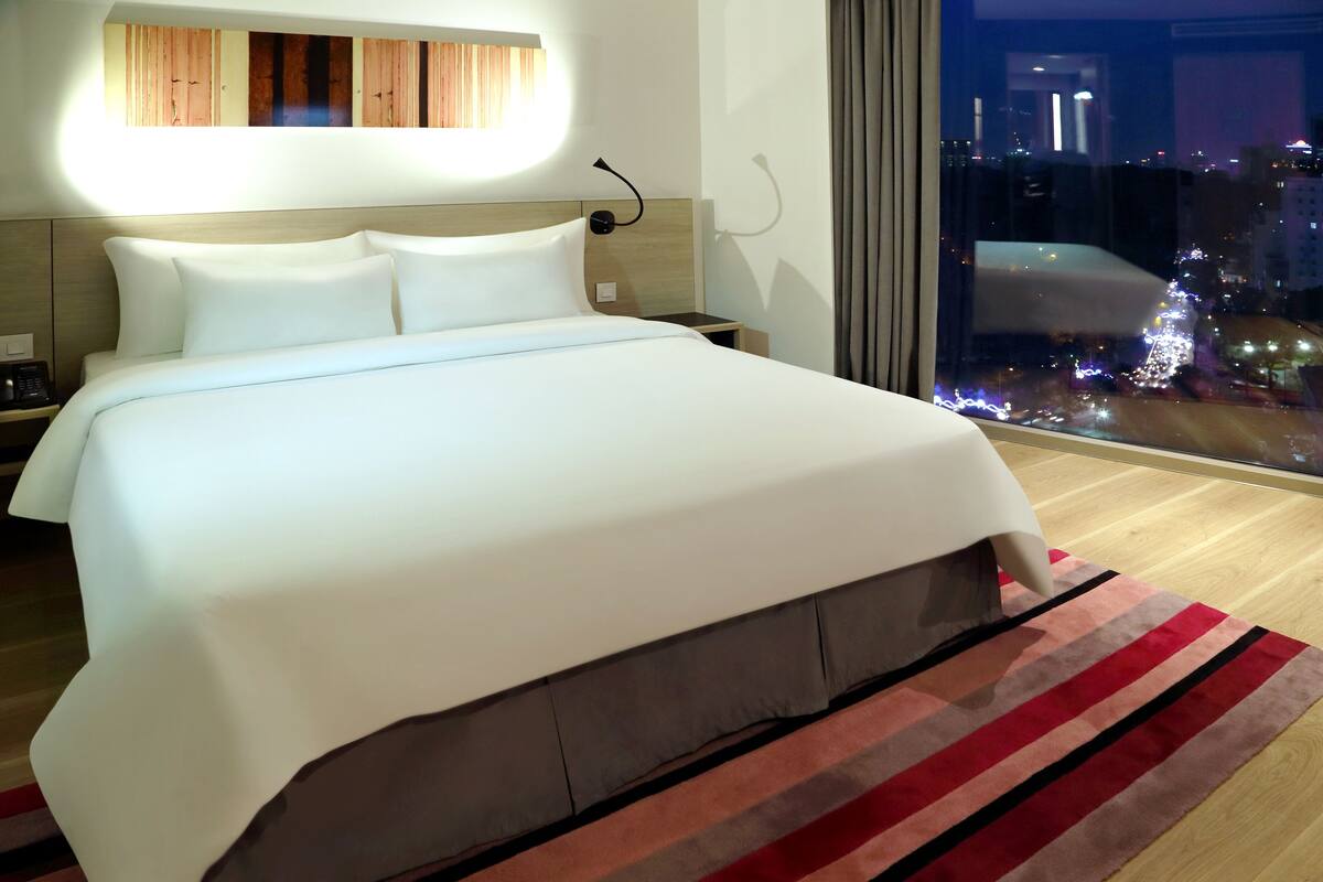 Ein modernes Hotelzimmer mit einem großen Bett, einem Nachttisch, einer Wandlampe und Blick auf die beleuchtete Stadt bei Nacht.