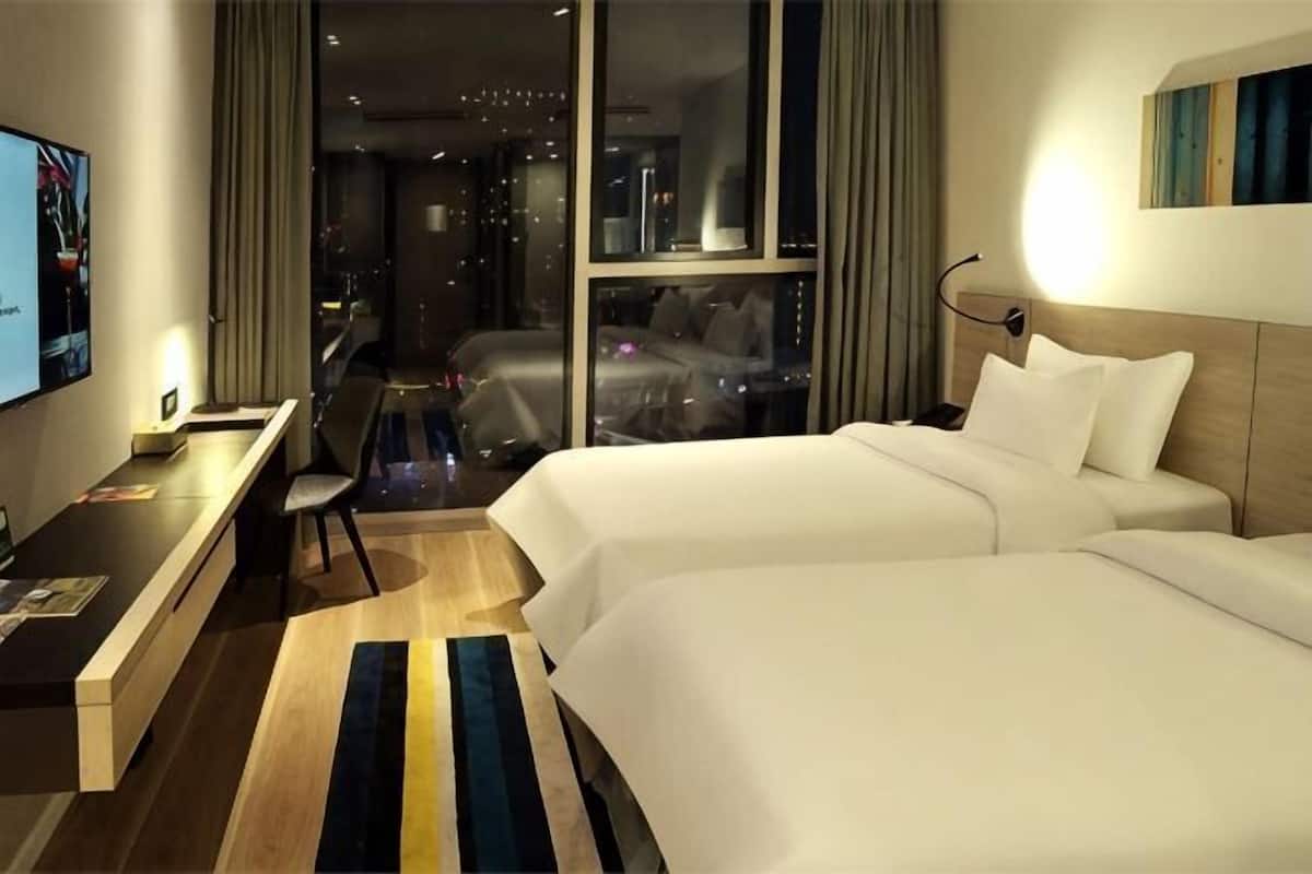Ein Hotelzimmer mit zwei Betten, einem Schreibtisch, einem Sessel, einem Fernseher und Blick auf die Stadt.