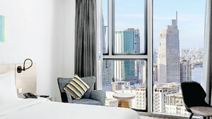 Hotellihuone, jossa on suuri sänky, työpöytä, tuoli ja suuret ikkunat, joista avautuu näkymä kaupungin ylle.
