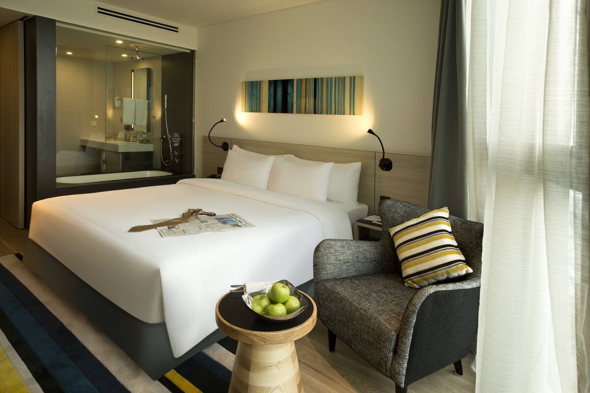 Ein Hotelzimmer mit einem großen Bett, einem Nachttisch mit einer Schüssel Äpfeln, einem grauen Sessel, einem modernen Badezimmer mit einem Spiegel und einem Wandbild.