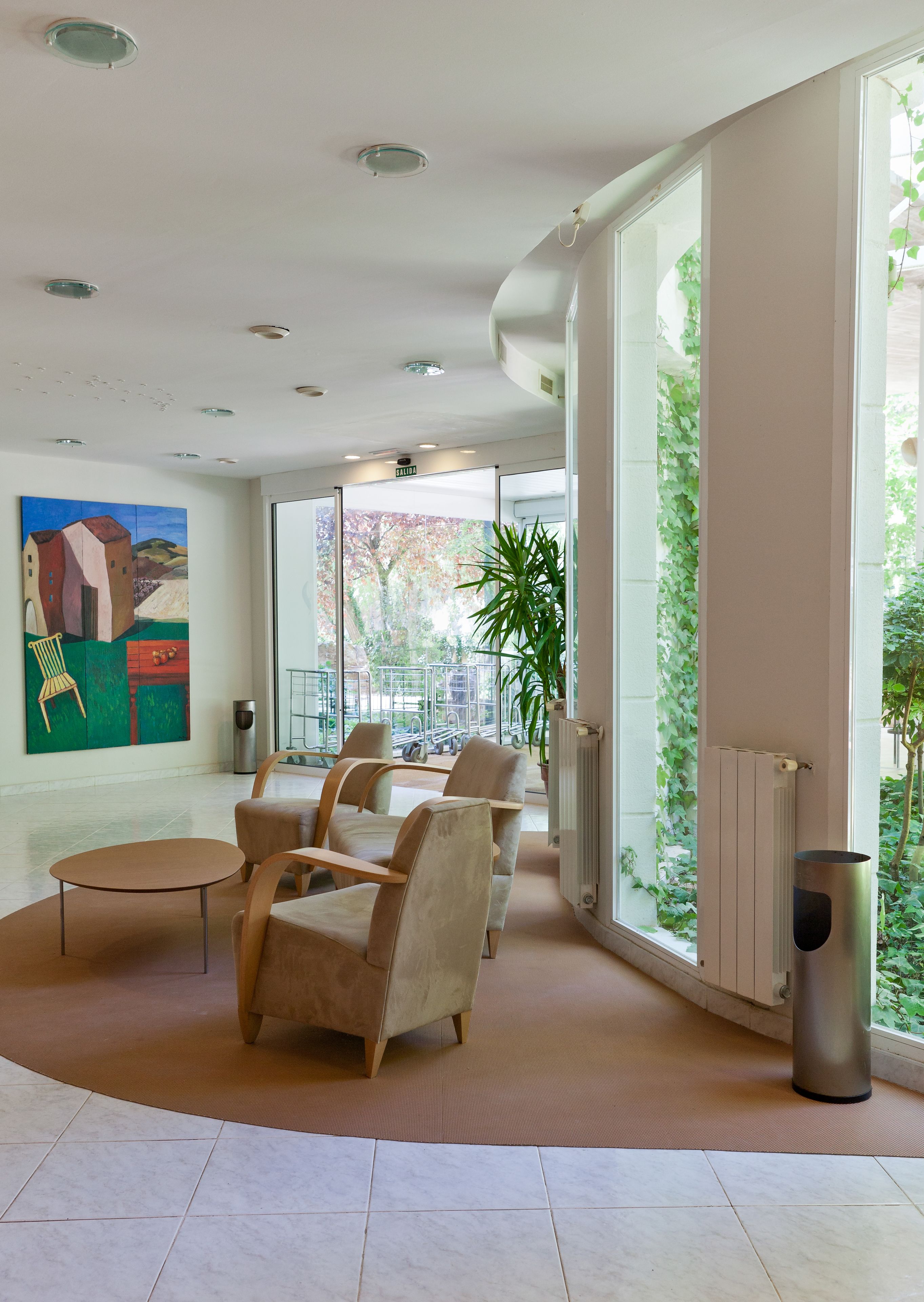 A spacious lobby with a modern design, featuring a seating area with beige chairs and a small table, a large window with a view of greenery, and a painting on the wall.