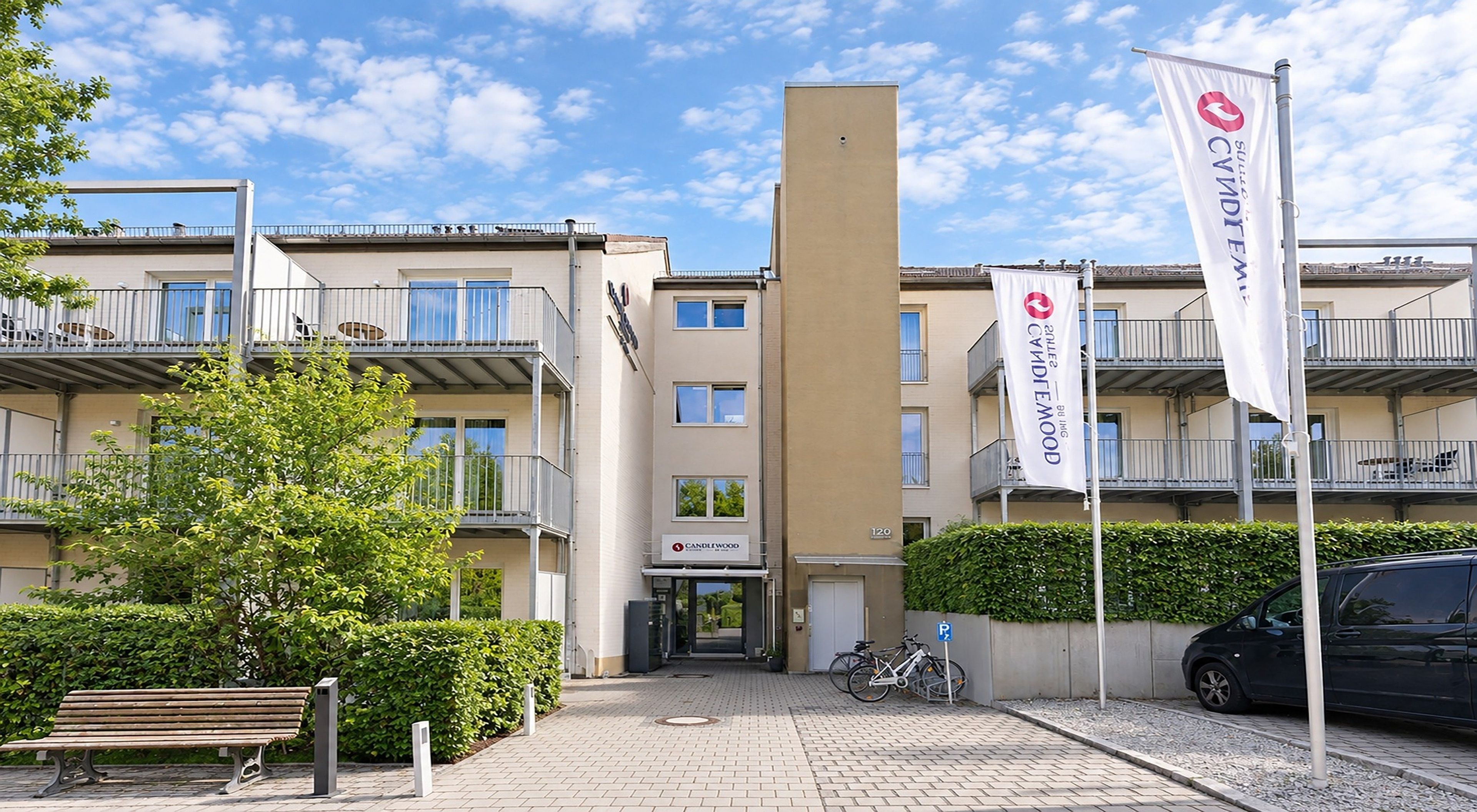 Un immeuble à plusieurs étages doté de balcons, un vélo garé à l’extérieur et un banc au premier plan.